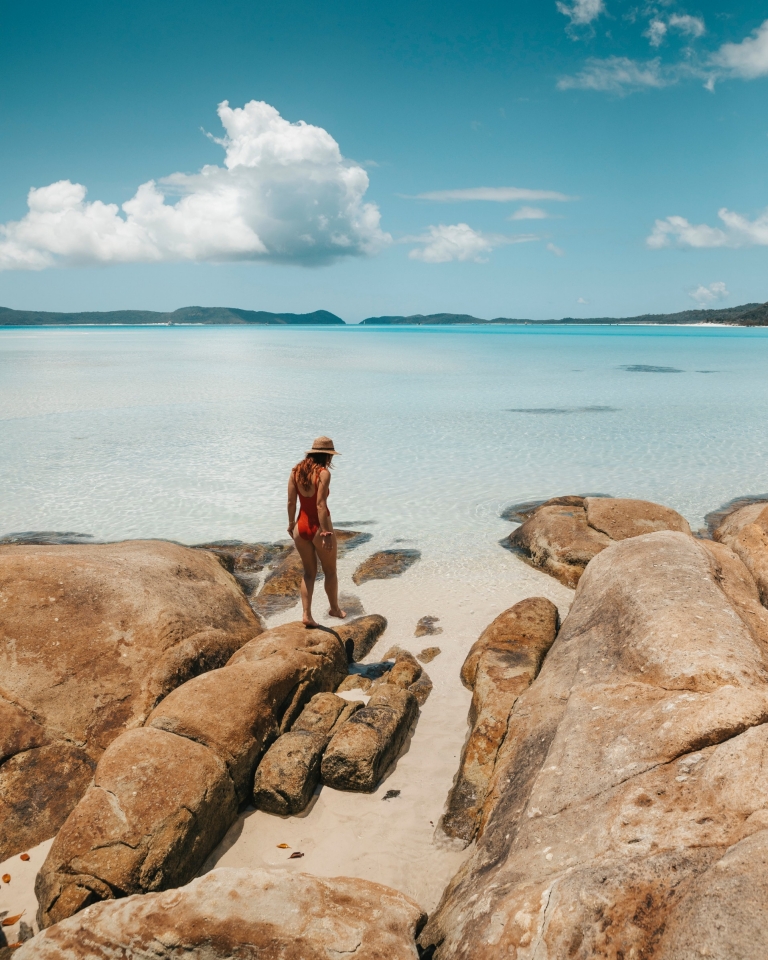 A woman in a red swimsuit walks on rocks at the ocean's edge at Whitehaven Beach, Whitsundays, Queensland © Tourism and Events Queensland